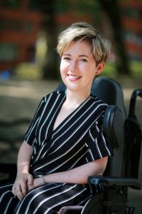 Image of Sarah Rennie, a white woman with short blonde hair sitting in her powerchair smiling towards the camera