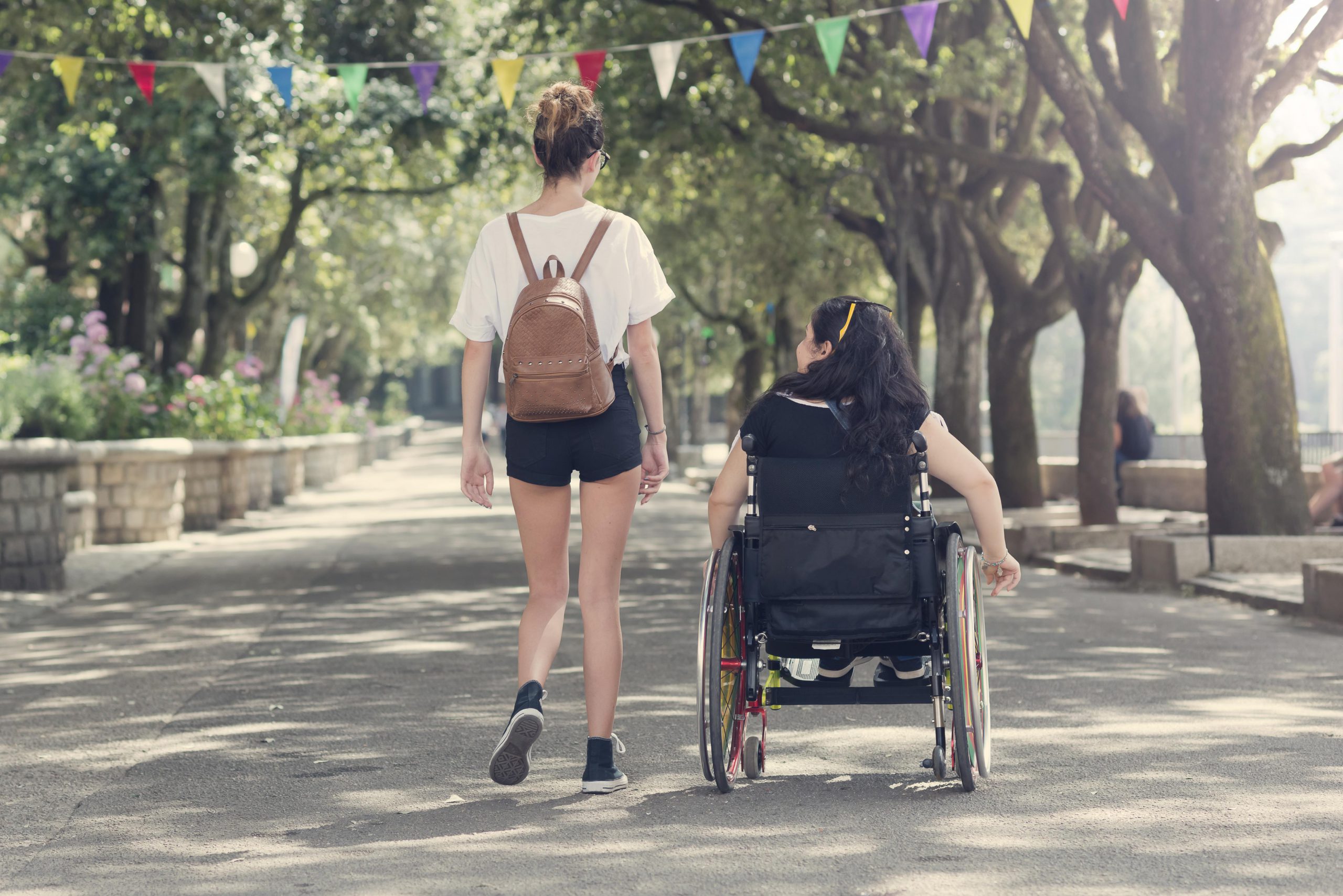 One woman who is walking and another who is steering her wheelchair travel down a tree-lined path side by side