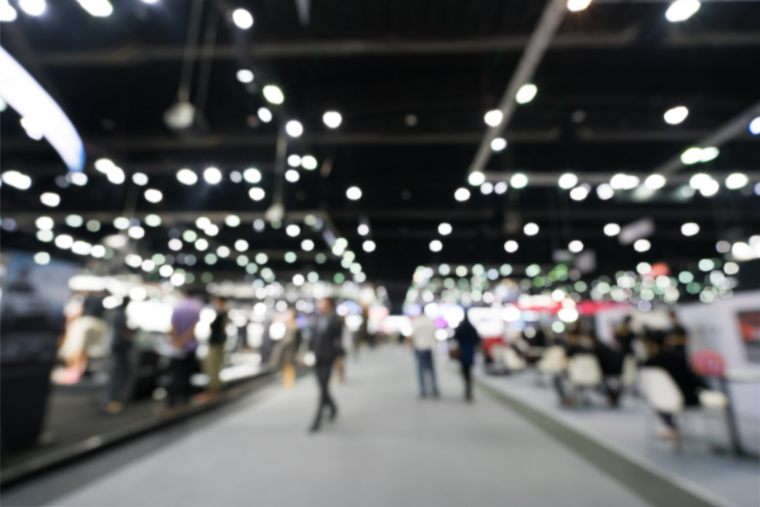 A blurred image of Naidex conference space where we see outlines of people, stalls and lights