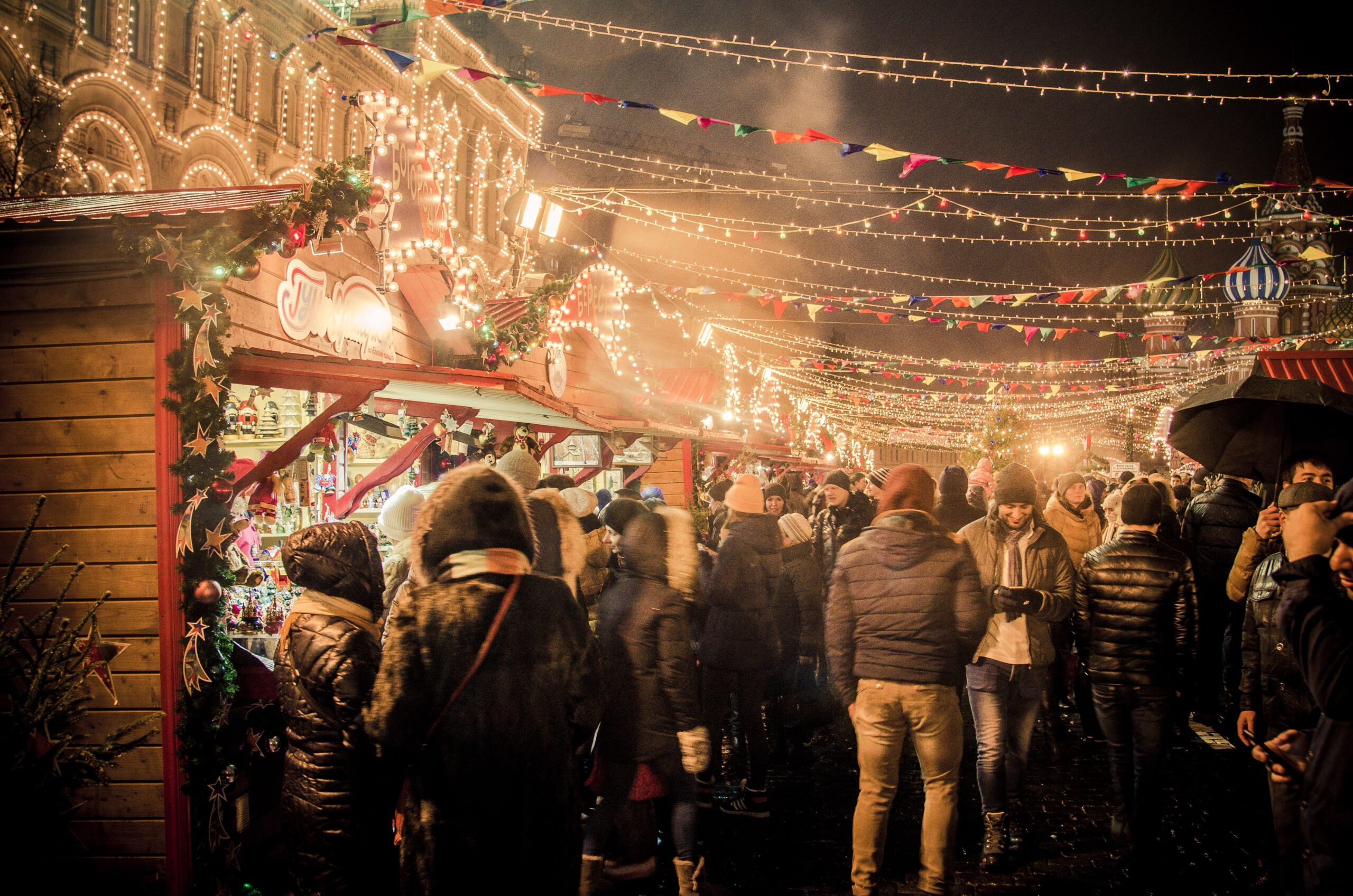 Photo of a Christmas market with lots of different food stalls and a crowd of people wearing jumpers and coats standing around them. The image is dark and cosy.