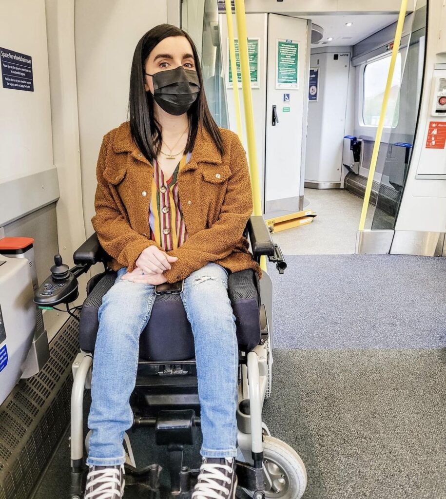 Emma is in her wheelchair on a train travelling. She is wearing a brown jacket, blue jeans and converses. 
