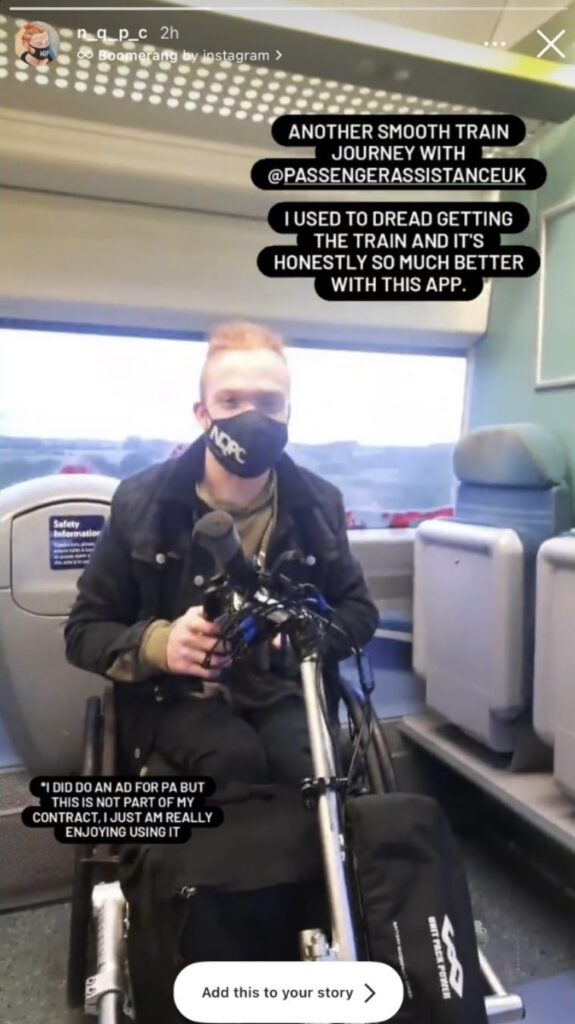 Charlie, a ginger haired male, is on his  wheelchair on a train. He is wearing a black face mask, a black jacket and a green t-shirt.