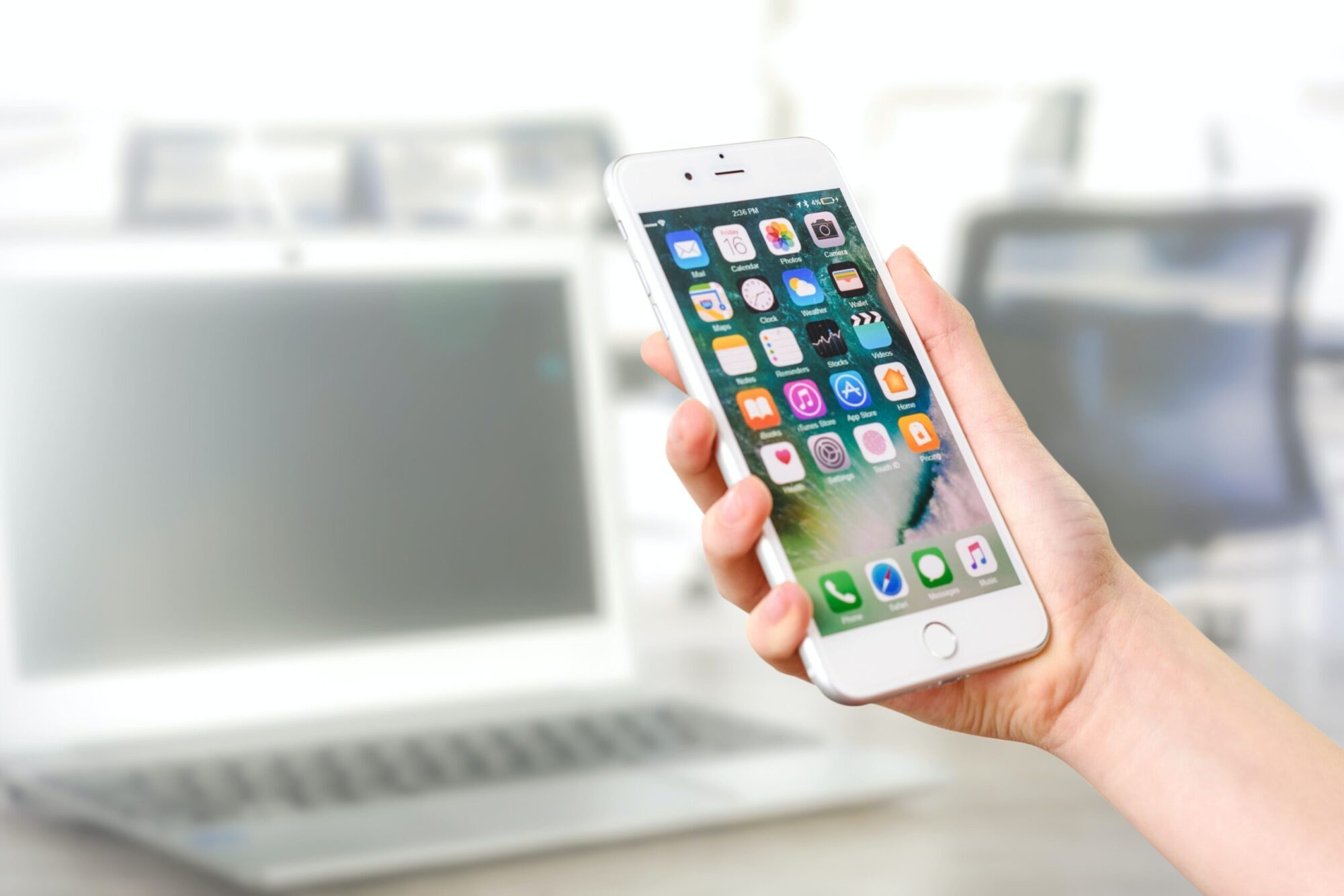 Photo of a male holding his iphone showing the home screen. His white laptop is in the background.