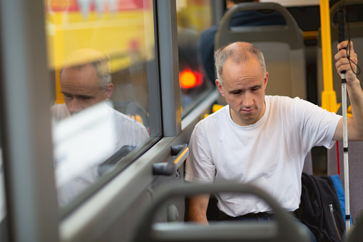 adult blind man sitting in bus, wearing white shirt and jeans, one hand at his blind persons cane