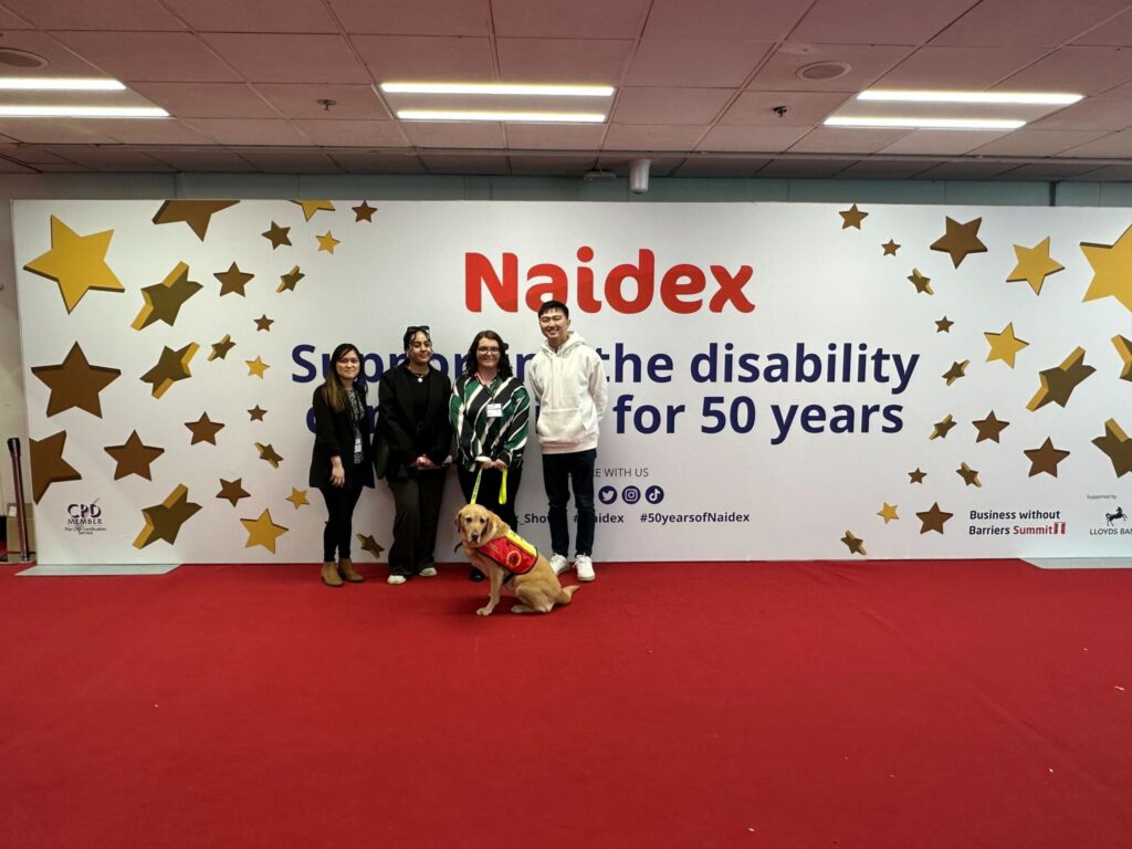 4 people and a assistance dog facing and smiling at the camera in front of a Naidex banner