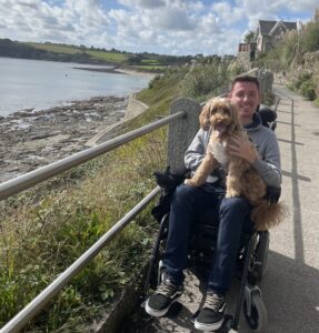 Ross Lannon, a caucasian man in a power chair, holding his dog Ralph. They are on a costal path. There is a grassy bank leading to a beach on the left hand side of the image. There is a house and hills in the background.