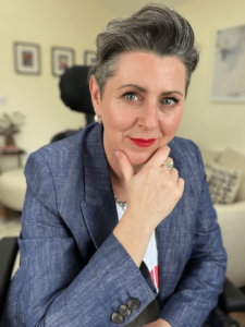 Sandie Roberts has short grey hair and red lipstick, wearing a blue/grey blazer. Sandie sits holding their chin in one hand. 