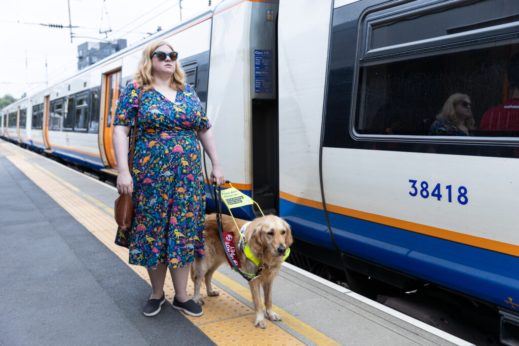 Dr Amy Kavanagh, a white blonde woman wearing a colourful dress and black glasses, standing beside her guide dog on a train station platform with a train to their side and standing on tactile paving.