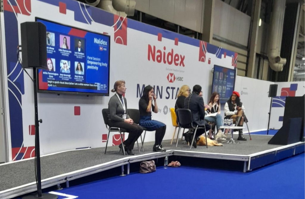 6 people and an assistance dog on a grey stage. To their left, 2 BSL interpreters are also seated. There are 1 screens showing images of the panel on the wall behind them, which also has the "Naidex main stage" sign.