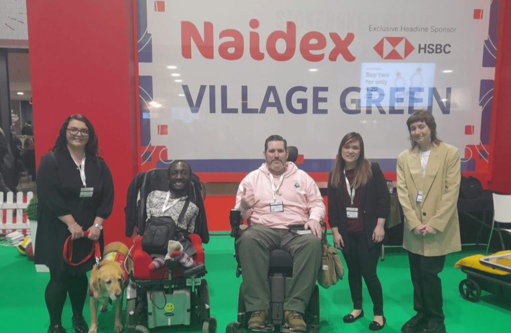 5 people on a green floor in front of a red and white background that says "Naidex village green". From left to right: Emma Partlow, a caucasian woman with long dark hair and glasses, holding an assistance dog on a red lead. Isaac Harvey, a black man in a patterned shirt, smiling in a power chair. Nick Wilson, a caucasian man in a pink hoodie, smiling in a power chair. Linda Truong, a south east asian woman with long brown hair wearing a black suit. Mary Senier, a caucasian woman with short brown hair in a tan blazer.