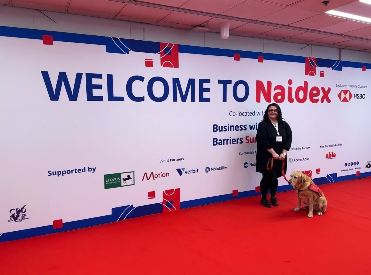Emma Partlow, a caucasian woman with long dark hair and glasses standing on a red carpet in front of the "Welcome to Naidex" sign. Emma is smiling and holding her assistance dog Luna on a red lead.