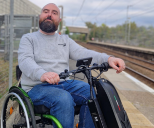 Image of Joe, a white man with a shaved head and short auburn beard, smiling. Joe is wearing blue jeans and a grey sweatshirt and sits in his green wheelchair at a small train station.