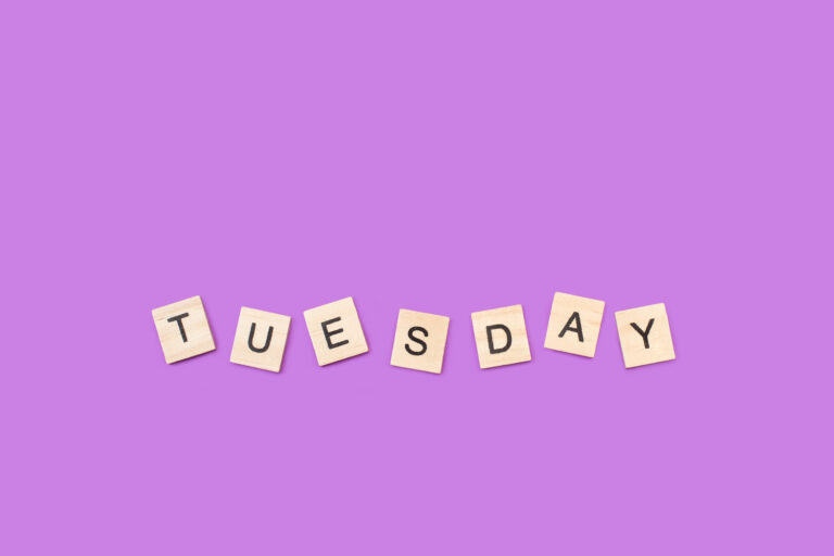 Wooden scrabble tiles on a purple background spell out "Tuesday".