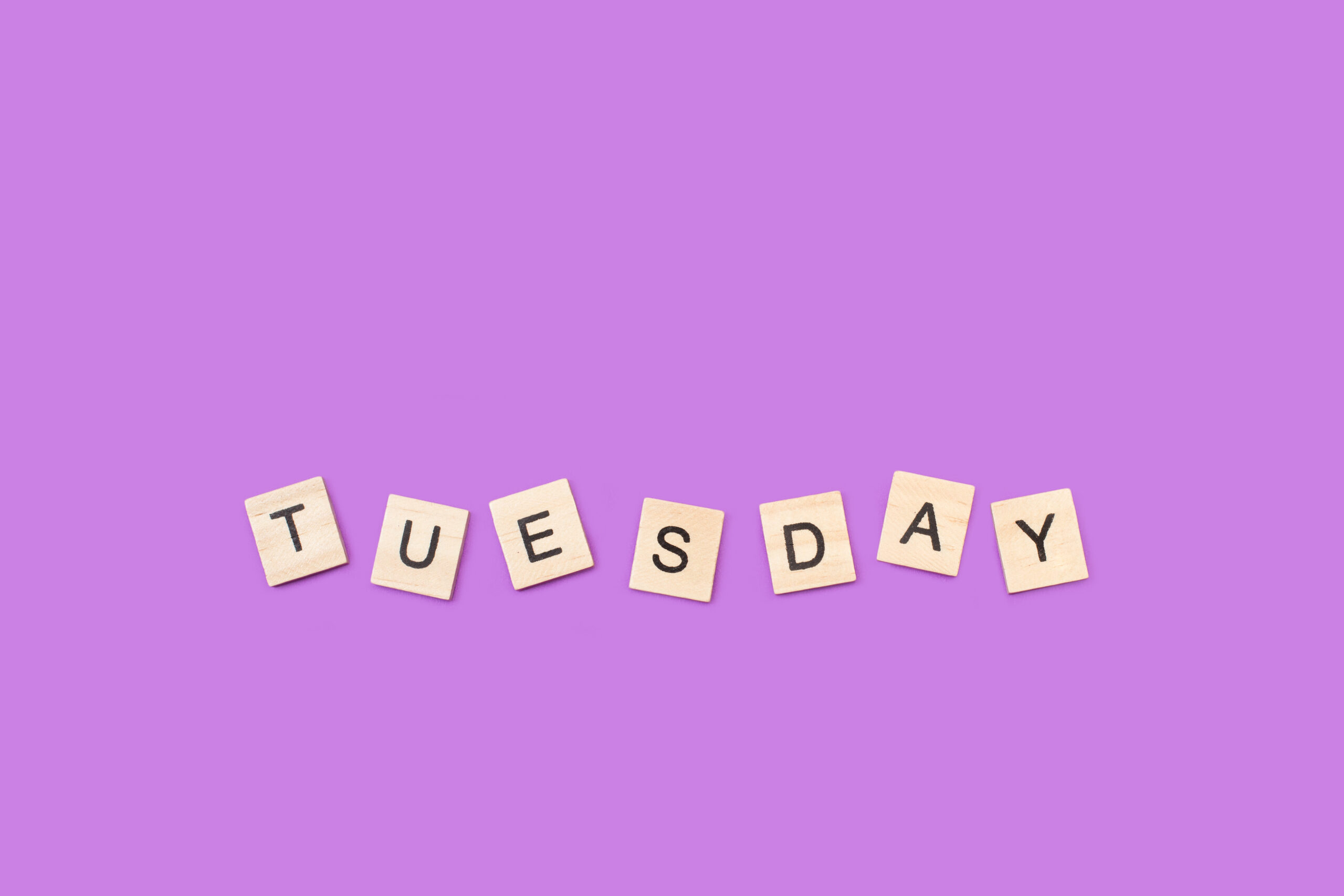 Wooden scrabble tiles on a purple background spell out "Tuesday".