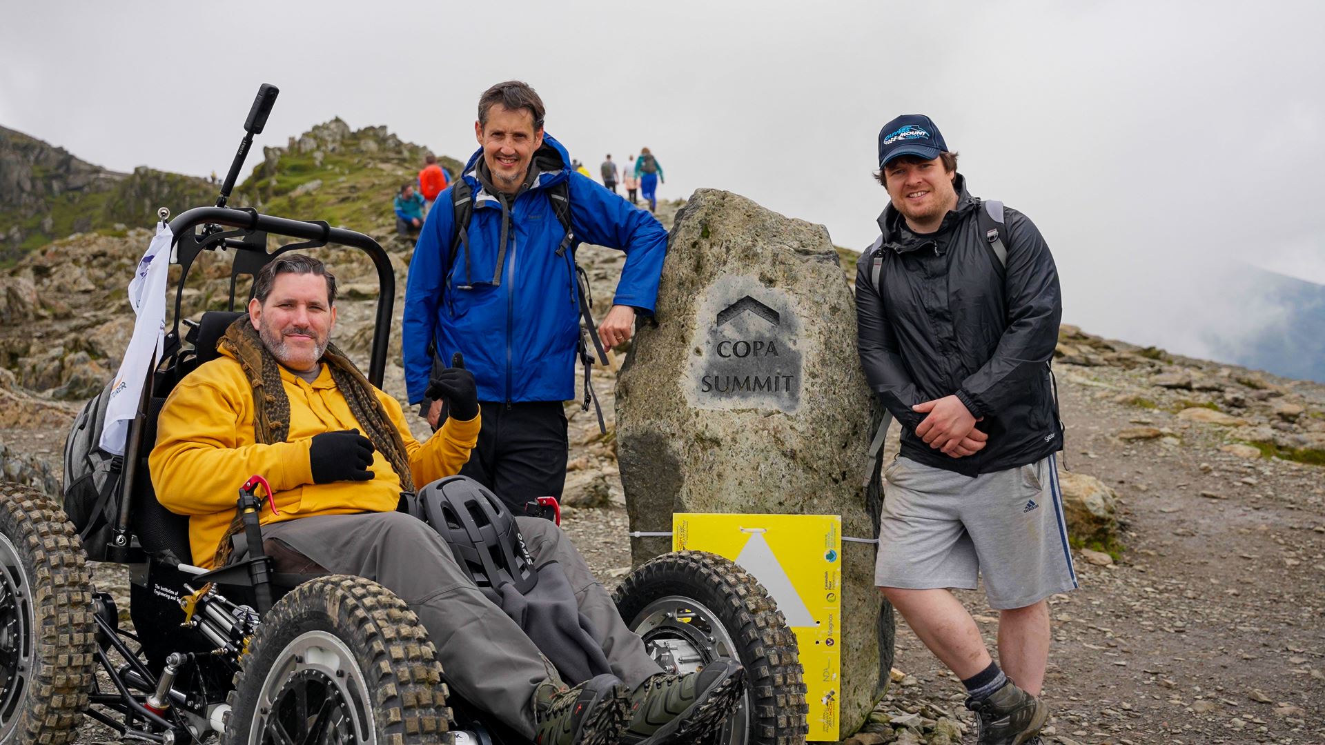 Nick Wilson in RockClimber beside the Copa Summit sign on Mount Snowdon. Nick is smiling, wearing a yellow hoodie, grey trousers, and black gloves.