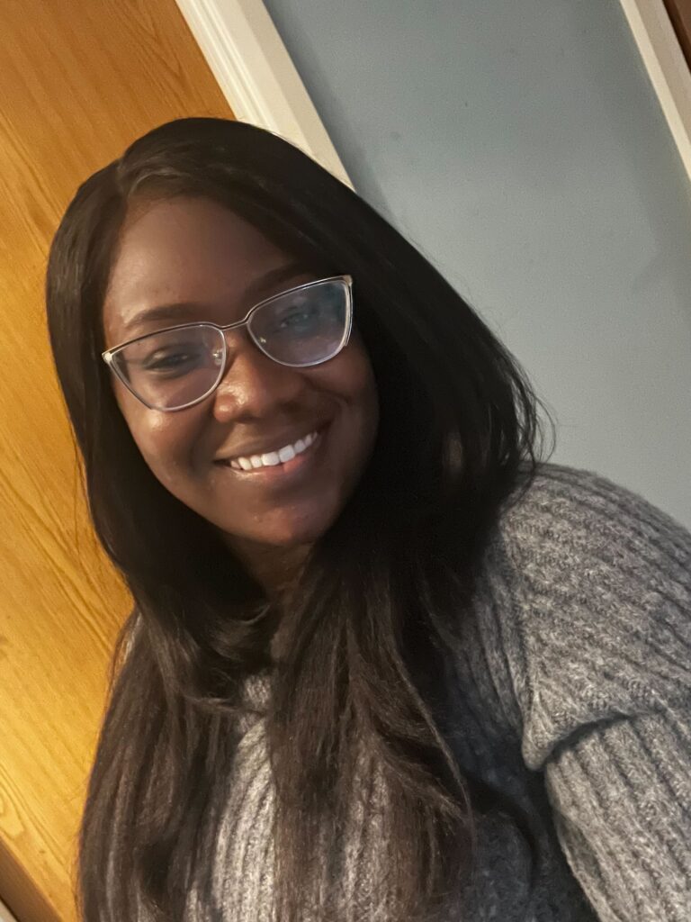 Becky smiling at the camera indoors, wearing glasses and a grey jumper, with long dark hair and a wooden door in the background.