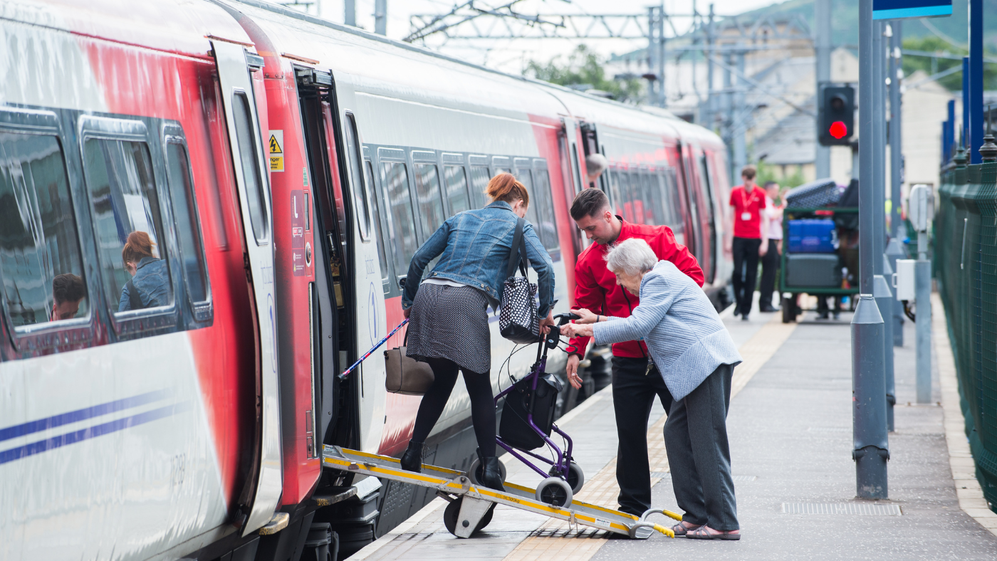 Rail staff using a ramp to support an older passenger with a mobility aid boarding a train.
