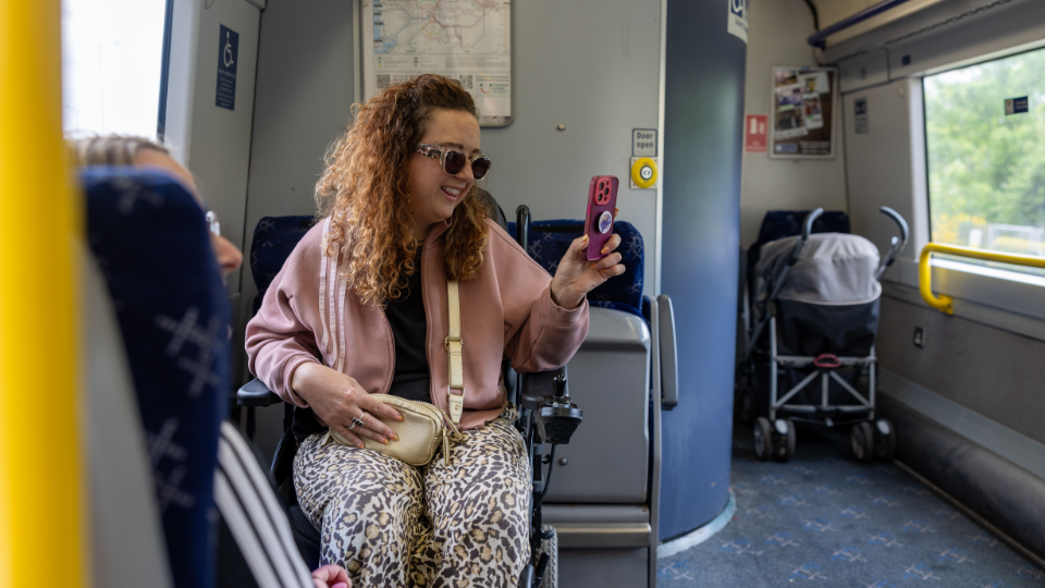 Carrie Ann Lightley smiling and looking at her phone while travelling in the wheelchair space on a train.