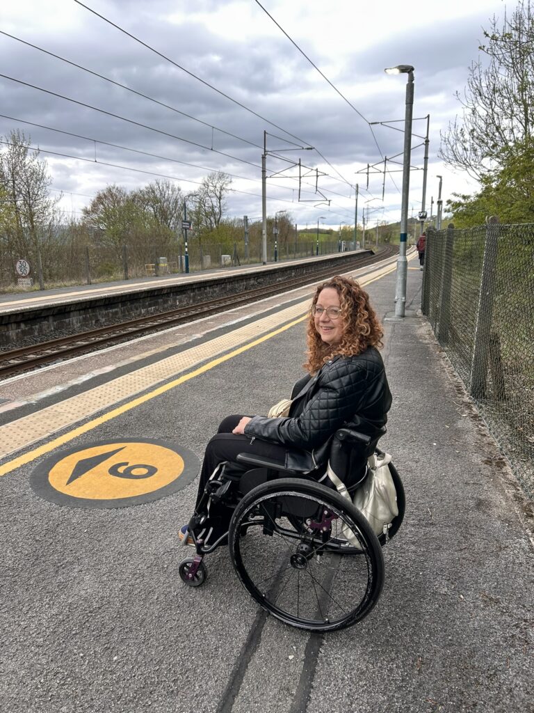 Carrie Ann Lightley smiling while waiting in her wheelchair on a train station platform.