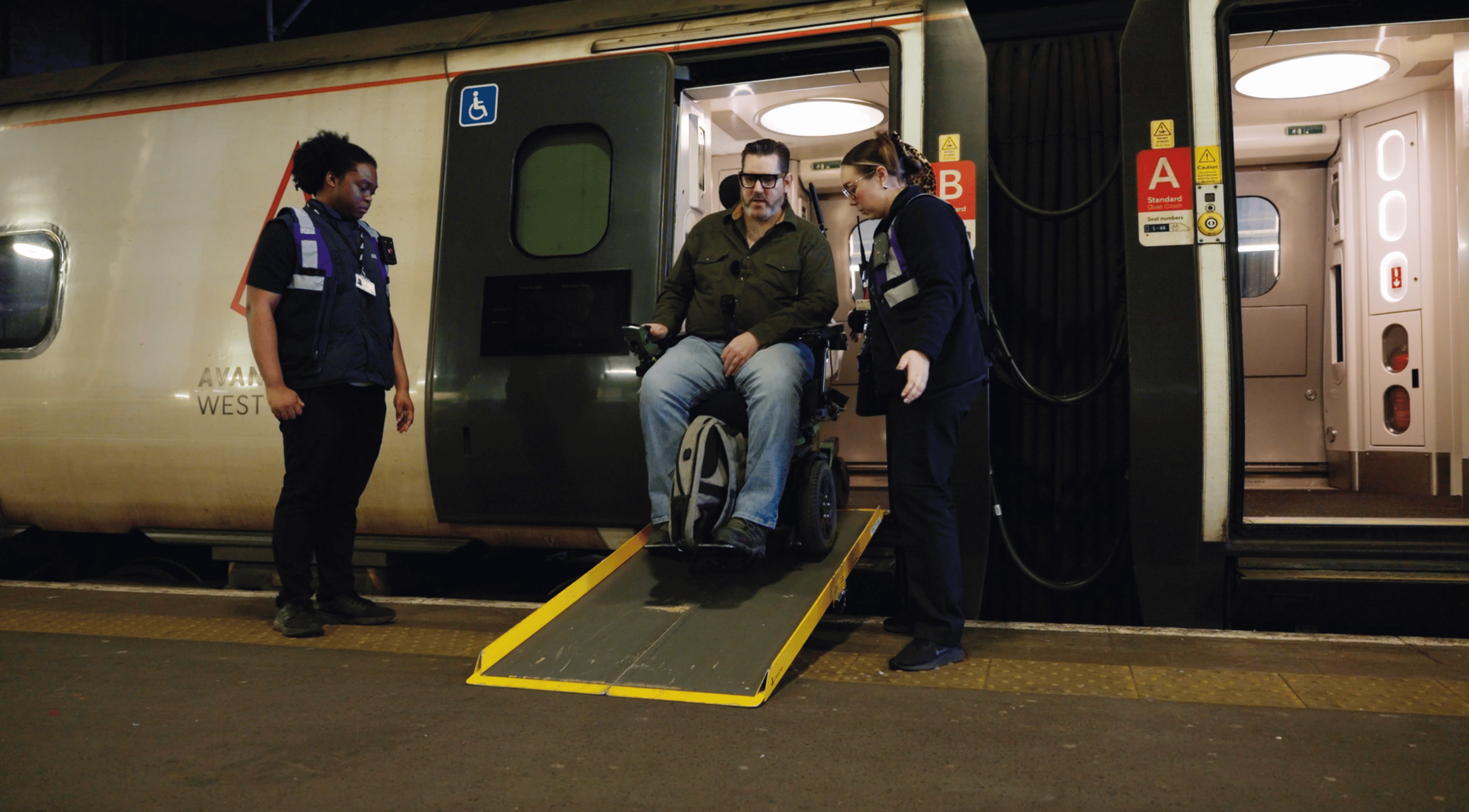 A wheelchair user getting off a train using a boarding ramp, with station staff providing assistance.