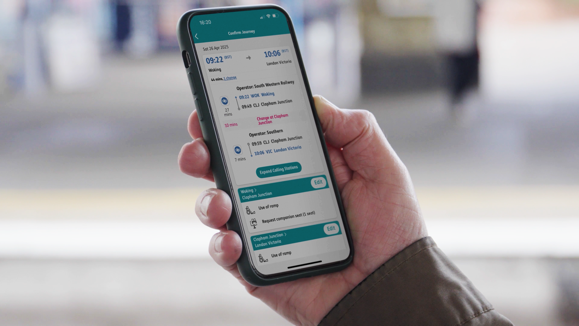 A hand holding a phone displaying the ‘Confirm Journey’ page in the Passenger Assistance app while standing on a train platform.