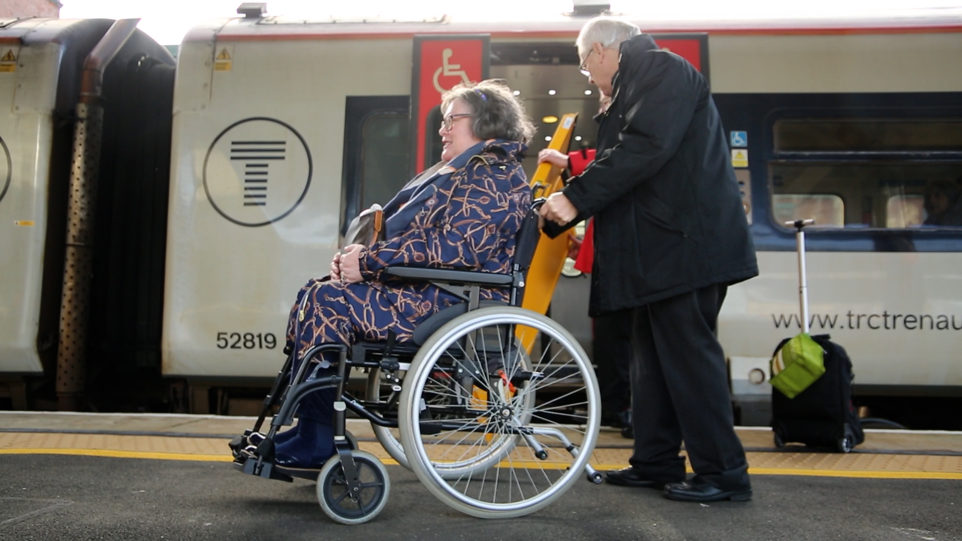 A wheelchair user is supported by a station staff on the train platform.