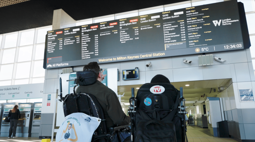 Two wheelchair users looking at the journey details on the information board at the station