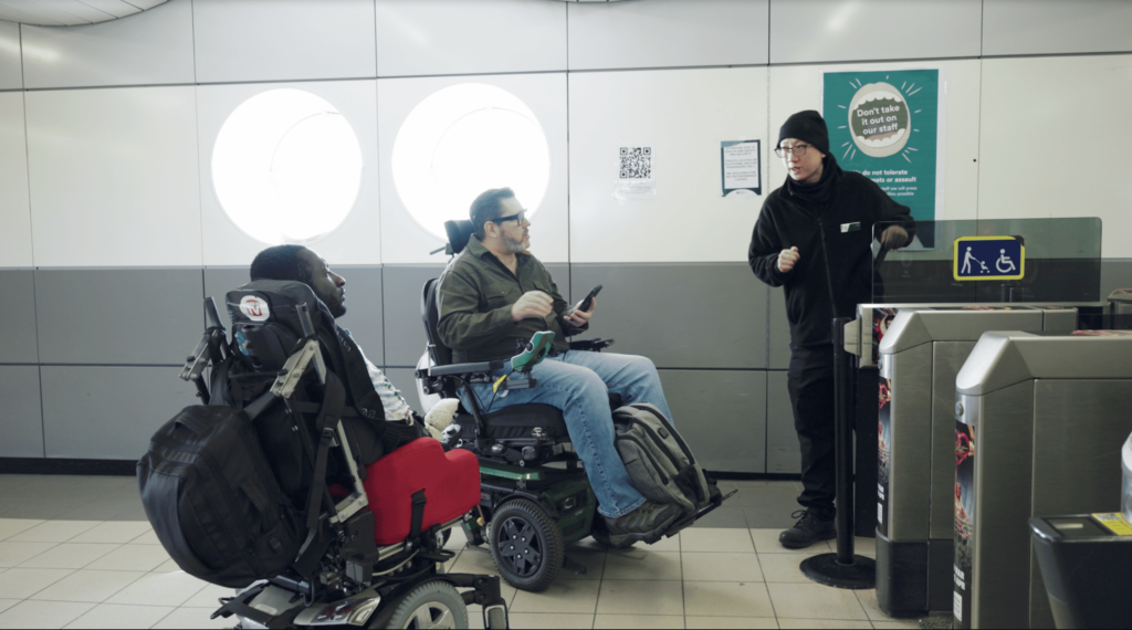 Two wheelchair users talking to a station staff at the ticket barriers