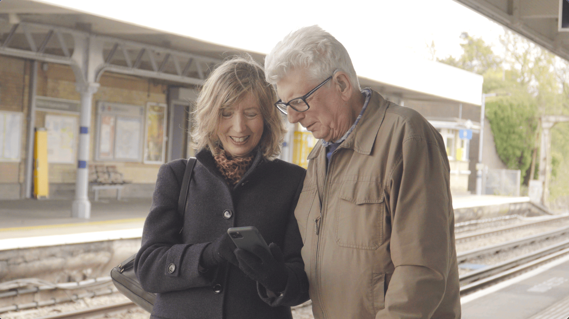 An elderly couple smiling as they look at the phone screen on the platform of a train station.